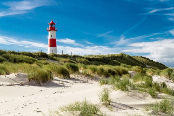 Rot weisser Leuchtturm steht in sanften Sanddünen mit Strandgras unter blauem Himmel an der Nordseeküste