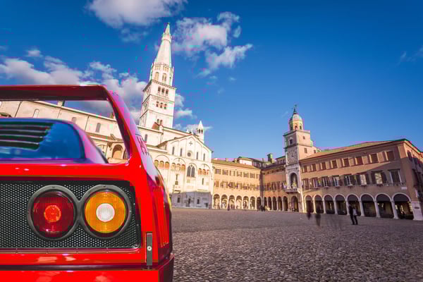 Historischer Domplatz in Modena mit romanischem Dom und Glockenturm unter blauem Himmel, im Vordergrund das Heck eines roten Sportwagens