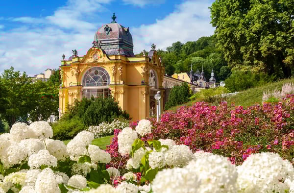 Blumenreicher Kurpark in Marienbad mit Blick auf ein historisches Kurgebäude und bewaldete Hügel