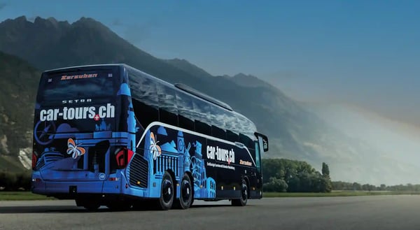 Reisebus steht auf einer grossen Asphaltfläche zwischen grünen Wiesen und hohen Bergen unter hellem Himmel