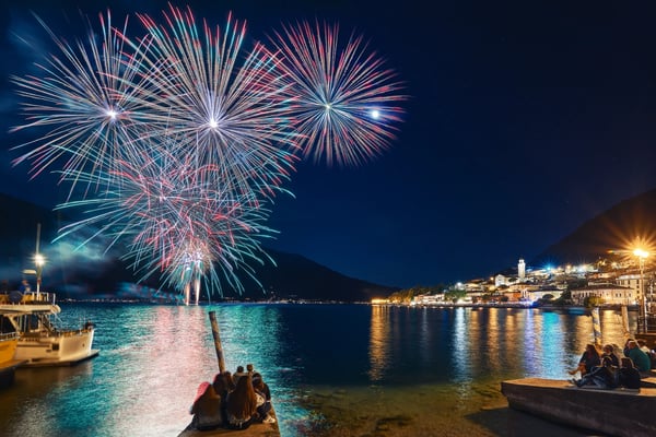Buntes Feuerwerk über dem Gardasee bei Nacht mit Spiegelung im Wasser und beleuchteter Uferstadt