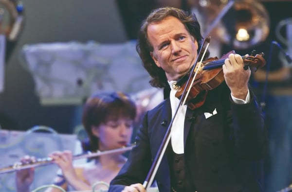 Violinist in elegantem Anzug spielt auf der Bühne begleitet von einem Orchester im Hintergrund.